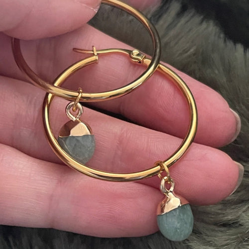 Gold hoop earrings with green stone charms held in a hand against a fur background