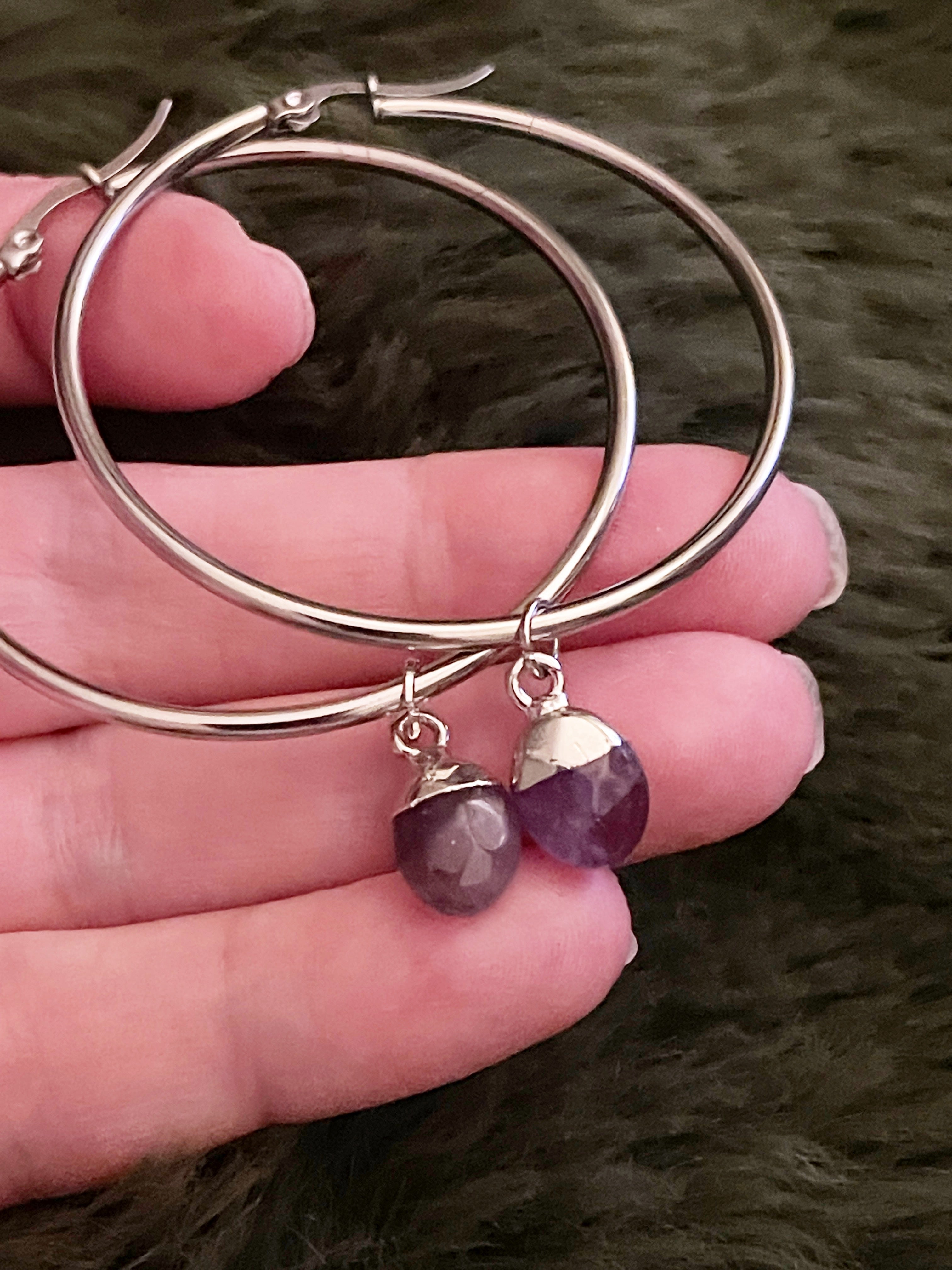 Silver hoop earrings with purple gemstone crystals held in a hand against a dark background