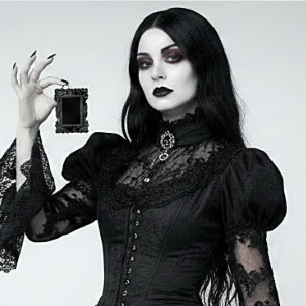 A woman with long black hair and gothic makeup wears a black lace and corset-style dress while holding a small black rectangular picture frame against a plain light grey background.