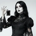 A woman with long black hair and gothic makeup wears a black lace and corset-style dress while holding a small black rectangular picture frame against a plain light grey background.