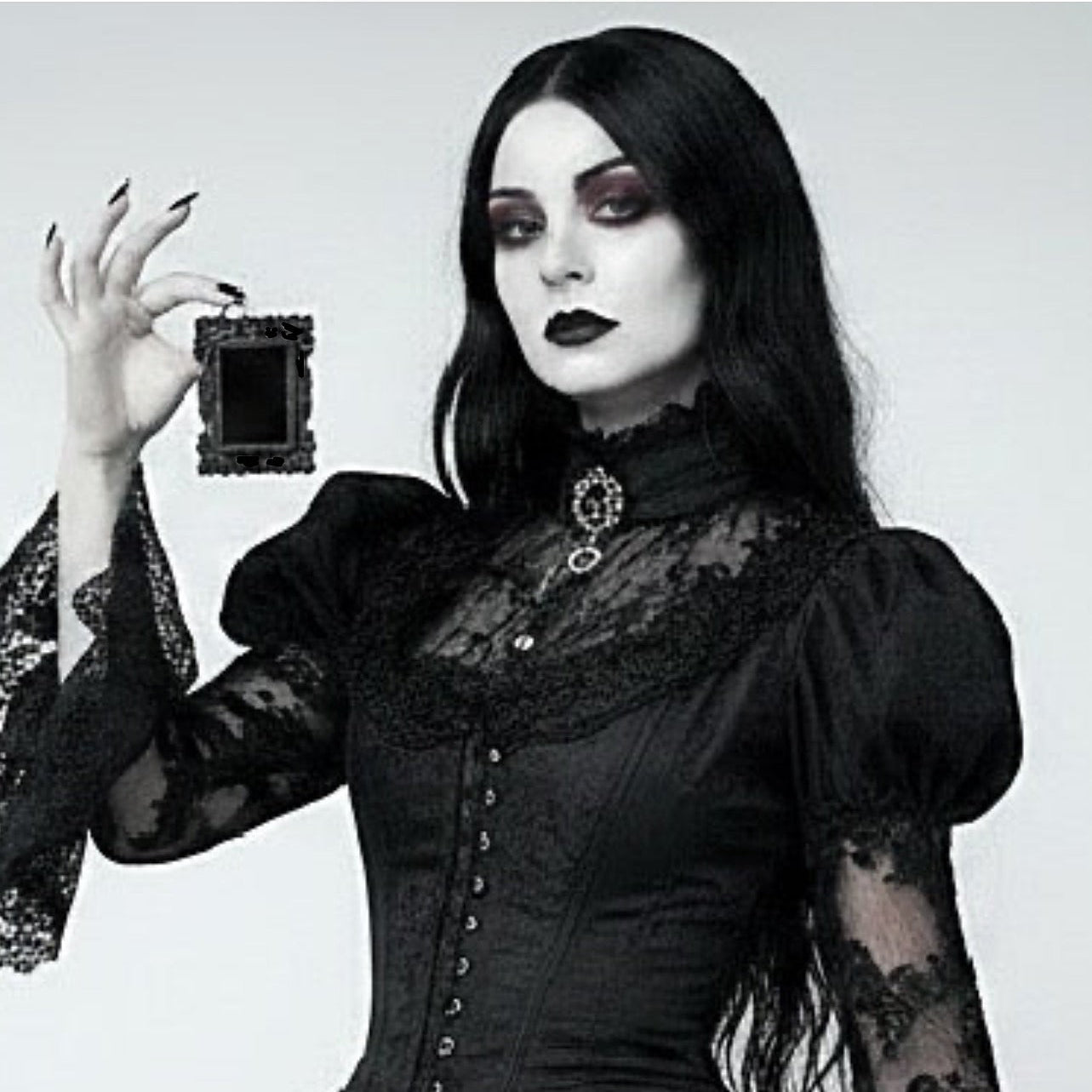 A woman with long black hair and gothic makeup wears a black lace and corset-style dress while holding a small black rectangular picture frame against a plain light grey background.